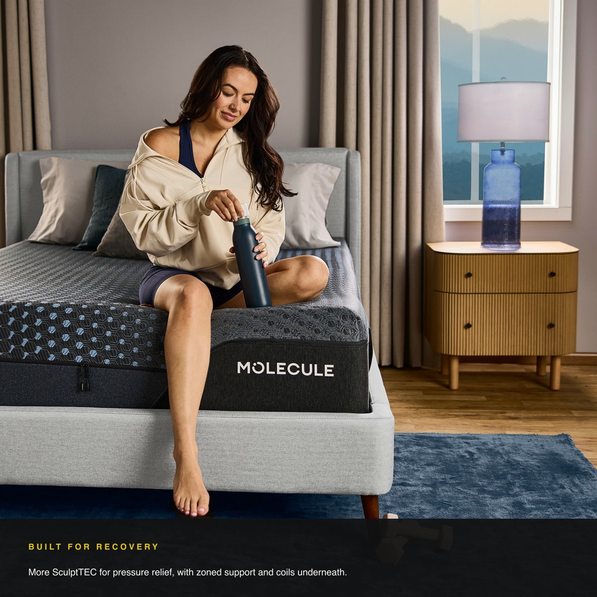 Woman sitting on the edge of the MOLECULE Engage Pro Hybrid 13 mattress with a water bottle