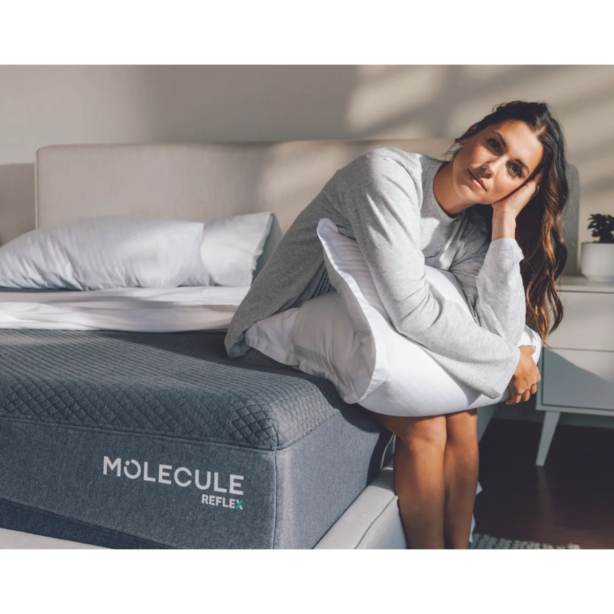 A person sitting on a bed with a gray MOLECULE Reflex mattress in a bright bedroom.